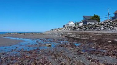 Bulutsuz bir denizin ve açık mavi bir gökyüzünün altında kıyıda birkaç evi olan kayalık bir sahil sahilinde alçak uçuşlar. Saint-Ulric-de-Matane, Quebec, Kanada, 2025.
