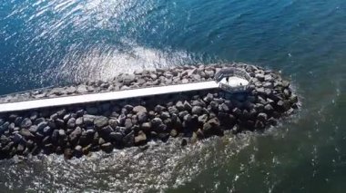 Tahta bir tahta kaldırım ve mavi su dalgalarıyla çevrili küçük bir gözlem platformu ile koruyucu bir dalga kırığı oluşturan taştan bir dalganın hava görüntüsü. Bonaventure, Quebec, Kanada, 2025.