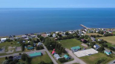 Ufukta gökyüzüne karışan sakin mavi okyanusun yakınındaki eski bir rıhtım, renkli evler, yeşil parklar ve spor sahalarını gösteren bir kıyı köyünün havadan görünüşü. Baie-des-Sables, Quebec, Kanada, 2025.