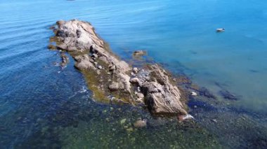 Kayalık bir resifin yakın mesafeden üstgeçişi, bir kayığın demirlendiği ve deniz kuşlarının dinlendiği sakin bir körfezdeki gelgitlerin ve havanın dokusunu aşındırdığını gösteriyor. Anse du Petit Mitis, St. Lawrence Nehri, Quebec, Kanada, 2025.