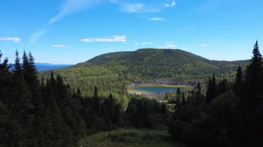 Yoğun ormanların panoramik hava manzarası, sakin bir göl ve yuvarlanan dağların arasından geçen dolambaçlı bir yolun yakınındaki odun yığınları. Sainte-Irene, Quebec, Kanada, 2025.