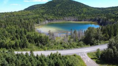 Yoğun bir ormana yerleşmiş turkuaz bir göl, kıyı boyunca dolambaçlı bir yol ve ufukta yuvarlanan dağlar. Joram Gölü, Sainte-Irne, Quebec, Kanada, 2025.