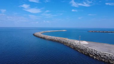 Uzun taştan bir dalgakıran sakin mavi denize doğru çıkar ve kıyı şeridi boyunca koruyucu bir bariyer oluşturur. Huzurlu bir kıyı manzarası, seyahat, limanlar veya denizle ilgili konular için ideal..