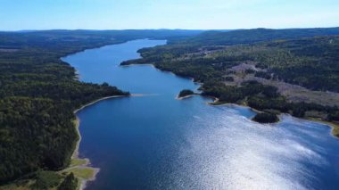 Bereketli ormanların ve yumuşak dağların arasından akan ve mavi gökyüzünün altındaki güneş ışığını yansıtan dolambaçlı bir gölün nefes kesici görüntüsü. Mitis Gölü, Quebec, Kanada, 2025.