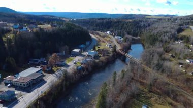 Sonbaharda paslı bir demiryolu köprüsü, yerleşim sokakları ve ormanlık tepeleri olan, kırsal bir köyde dolambaçlı, huzurlu bir nehir manzarası. Gasp Yarımadası, Causapscal, Quebec, Kanada, 2025.