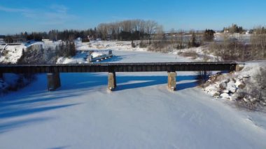 Demiryolu köprüsünün altından geçen insansız hava aracı videosu buzun üzerine dökülen uzun gölgeler ve huzurlu bir kırsal alanda donmuş bir nehri kapsıyor. Price, Quebec, Kanada, 2026.