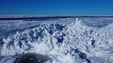 Buz dağları ve donmuş bir sahil boyunca uzanan karlar, açık mavi bir kış gökyüzü altında, küçük bir köy ve rüzgar türbinleri, St. Lawrence Nehri, Gaspe Yarımadası, Quebec, Kanada.