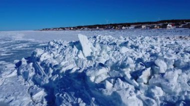 Donmuş deniz ve dik buz tepeleri bir kıyı şeridi boyunca birikiyor, açık mavi gökyüzünün altında rüzgar türbinlerinin bulunduğu küçük bir köy, Baie-des-Sables, St. Lawrence River, Quebec, Kanada, 2026.