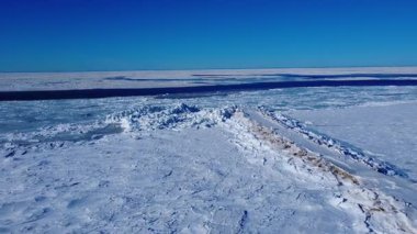 Ufuktaki açık mavi gökyüzünde kaybolan buzdan bir okyanusun kıyısında biriken buzun altında küçük bir iskele kapana kısılmıştır. St. Lawrence Nehri, Baie-des-Sables, Quebec, Kanada.