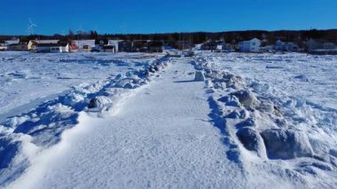 Buz kaplı eski bir rıhtım donmuş bir deniz boyunca uzanır. Açık mavi gökyüzünün altında küçük bir sahil köyüne doğru. Ormanın kenarında rüzgar türbinleri bulunur. Baie-des-Sables, Quebec, Kanada.