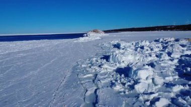 Donmuş bir deniz kıyısında uçarken mavi bir su kanalı bu kutup bölgesinden geçerek mavi bir gökyüzünün altında birkaç rüzgar türbini bulunan uzak bir sahil şeridine doğru yol alır. St. Lawrence Nehri, Quebec, Kanada.