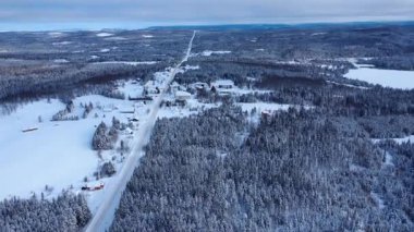 Kış havası, yoğun bir kozalaklı orman ve küçük bir köy boyunca kıvrılarak soğuk ve yalıtılmış kırsal yaşamı çağrıştıran karla kaplı kırsal bir yol gösteriyor. Sainte-Paule, Quebec, Kanada.