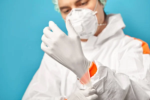 Nahaufnahme eines Arztes in Schutzkleidung, der sterile Handschuhe über schlichtem blauem Hintergrund anzieht. Umweltfreundlichkeit und Sicherheit bei medizinischen Eingriffen. — Stockfoto