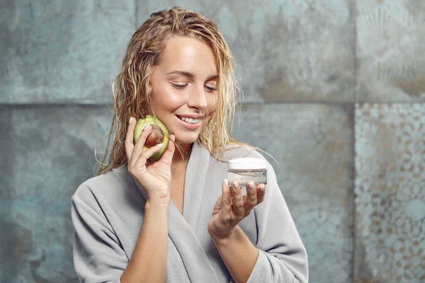 Attractive blonde hair woman after shower hold face cream and piece of avocado sitting in the bathroom, going to apply the moisturizer, has a happiness face expression.