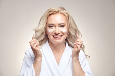 Gülümseyen orta yaşlı beyaz bornozlu bir kadın yüzüne krem sürüyor..