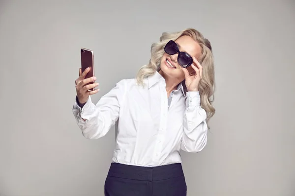 Mujer madura sonriente probándose gafas de sol y tomando selfies, retrato en gris aislado de señora mayor en un look casual con estilo —  Fotos de Stock