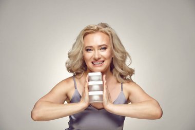 Gülümseyen yaşlı kadın gri izole olmuş vaziyette çeşitli krema kavanozlarını tutuyor.