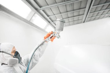 Odamda araba boyama elementlerini boyayan erkek bir tamirci. İşçi aracı sprey silahla beyaza boyuyor..