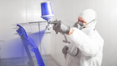 Araba boyacısı boya odasındaki araç parçasına mavi boya püskürtüyor..