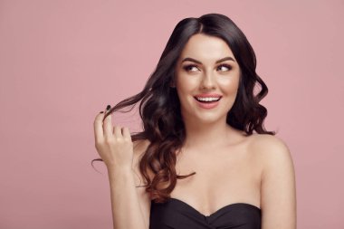 Çekici beyaz kadın samimi ve ilgili bir bakışla pembe izole bir arka plana bakıyor, kumral dalgalı saçlarına dokunuyor, kahverengi gözleri var, zevk veriyor ve şaşırtıyor..