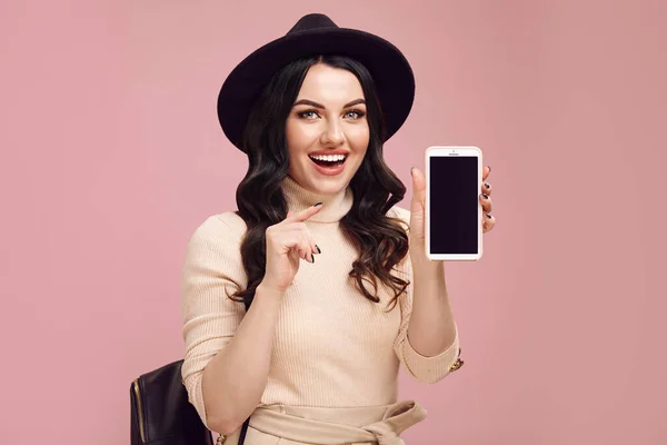 Chica adulta alegre señala a la pantalla en blanco de su teléfono y sonríe sorprendido mientras está de pie sobre rosa aislado —  Fotos de Stock