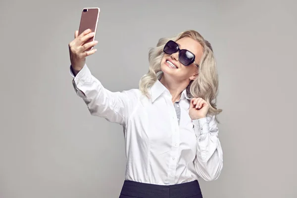 Mujer madura feliz en gafas de sol toma una selfie para compartir en las redes sociales en gris aislado —  Fotos de Stock