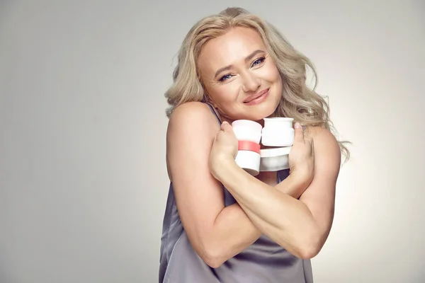 Adult fifty years old woman with clean skin cuddles with love to chest few various jars of moisturizer on a gray isolated background. Stock Image