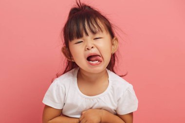İnatçı üç yaşındaki Asyalı küçük kız kaprisli bir şekilde pembe izole edilmiş arka planda dilini kameraya gösteriyor. Yaramaz çocuk konsepti, stüdyo resmi. 