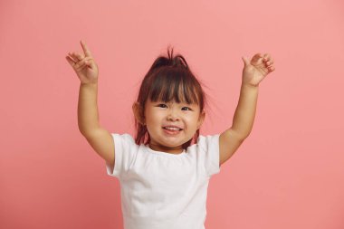 Neşeli üç yaşındaki Asyalı etnik kız ellerini kaldırıyor, neşeli bir ruh hali sergiliyor, pembe izole edilmiş bir tişörtte mutlu bir şekilde gülümsüyor.. 