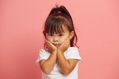 Sersemlemiş üç yaşındaki Asyalı kız ellerini pembe arka planda beyaz tişörtlü, yalnız bir şekilde durmaktan yanaklarına koyuyor. Şaşırmış bebek şok edici bir şeye inanamaz.. 