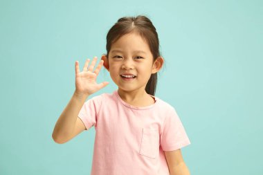 Tatlı Asyalı etnik kızın yatay portresi pembe tişörtlü neşeli bir şekilde el sallıyor kameraya gülümsüyor, pozitif bir havası var, mavi izole edilmiş arka planda boş duruyor.