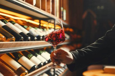 Şarap raflarının önünde tutulan kıvrımlı kırmızı şarap kadehinin yakın plan çekimi, vites hareketleri, kabarcıklar ve mahzen atmosferi.