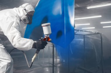 Koruyucu maskeli profesyonel sprey boyacı ve arabanın kaputunu boya odasında sprey tabanca kullanarak maviye boyayan kıyafetler. Boya kabininde araç boyama, restorasyon ve onarım..