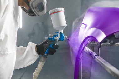 Araba boyacısı araba atölyesinde sprey tabanca kullanarak mor boyayı araba gövdesine püskürtüyor. Otomobil gövdesinin karmaşık restorasyonu, tam seçim rengi.