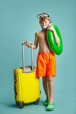 Stüdyo dikey portresi, turuncu yüzme şortu giyen, lastik daireli, su altı maskeli, sarı bavullu, mavi üzerine poz veren kırmızı saçlı küçük çocuk.