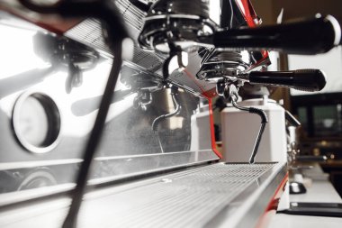 Yakın çekim profesyonel espresso makinesi, parlak iskele filtreleri ve buhar asasıyla. Makinelerin metalik yüzeyleri, kahvenin temizliğini ve hassasiyetini vurgulayan çevre ışığını yansıtıyor.