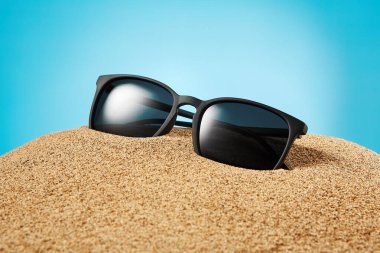 Güneş gözlükleri, siyah çerçeveli gölgeler, kısmen kumlara gömülü lensler mavi arka planda. Yaz mevsiminde, plajda, güneşli, turistik ve seyahatli. Yan görüş. Mesaj için yer. Boşluğu kopyala.