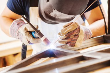 Yüz maskeli bir erkek, koruyucu eldiven kaynaklı argon-ark kaynaklı. Kaynakçı metal çerçeveye kaynak yapıyor. Tişört ve tulum giymiş bir işçi..