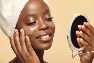 Güzel Afro-Amerikalı kadın göz maskesinin altına jöle bantlı temiz yüz derisi ile el aynası arıyor. Kırışıklıklardan, şişkinlikten, siyah halkalardan kadın kozmetik günlük rutini. Güzel cilt bakımı.