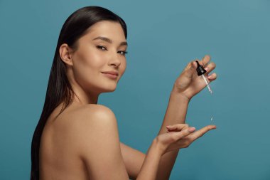 Asyalı kadın siyah kozmetik şişesini saklıyor, kameraya bakıyor. Bir damla serum, retinol, yağ damlayan pipet damlası tutan kızın parmağına damlıyor. Çıplak omuzlu güzel bayan, tekrar mavi. Cilt bakımı.