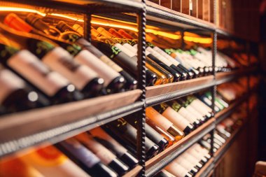 Aydınlatılmış şarap rafları temiz cam şişe çeşitliliği sunar. Kırmızı, beyaz ve güller dahil. Endüstriyel ahşap raflar görünür..