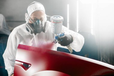 Araba boyacısı atölye garajında sprey tabanca kullanarak aracın bir kısmına kırmızı boya püskürtür. Boya odasında çalışan adam. Karmaşık onarım ve restorasyon otomobili sonrasında tam renk seçimi