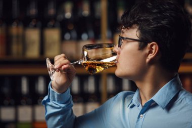 Asya kökenli şarap garsonu beyaz şarabı dikkatle tadıyor, şarap kalitesini değerlendirmek için elinde bardak tutuyor mahzende duran bulanık şişe arkaplanlarına karşı.. 