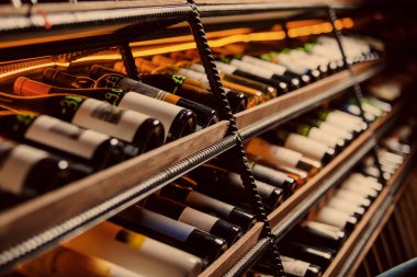 Ilık ışıklandırmanın altında ahşap raflarda istiflenmiş çeşitli şarap şişelerinin açılı görüntüsü, davetkar bir mahzen atmosferi yaratıyor..