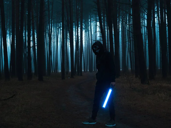Sisli, kasvetli bir ormanda parlayan neon ışığı olan maskeli bir adam. Seçici yumuşak odak, tonlu film