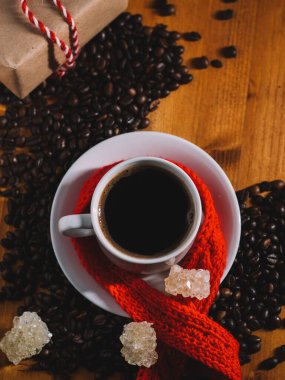 Beyaz espresso fincanı ahşap masanın üstünde kavrulmuş kahve çekirdekleri, şeker kristalleri, kırmızı sıcak eşarp ve sevgililer günü hediyesi.