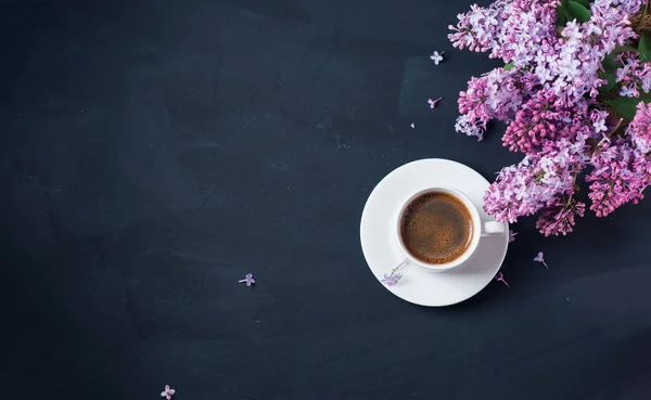 Koyu taştan bir masanın üzerinde beyaz bir fincan espresso. Mor leylak dalları, kopyalama alanı.