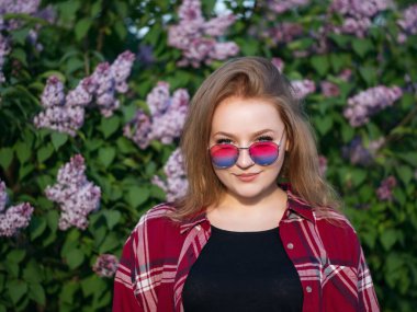 Renkli güneş gözlüklü çekici bir kızın portresi çiçek açan leylağın arka planında.