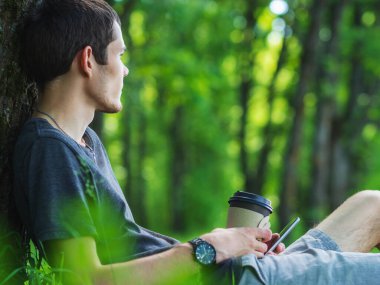 Genç bir adam parkta kahve içerek ve telefonla vakit geçiriyor. Açık hava eğlencesi