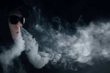 Tencere adam. Gözlüklü ve şapkalı genç beyaz bir adamın portresi elektronik bir sigaranın buharını boşaltıyor.